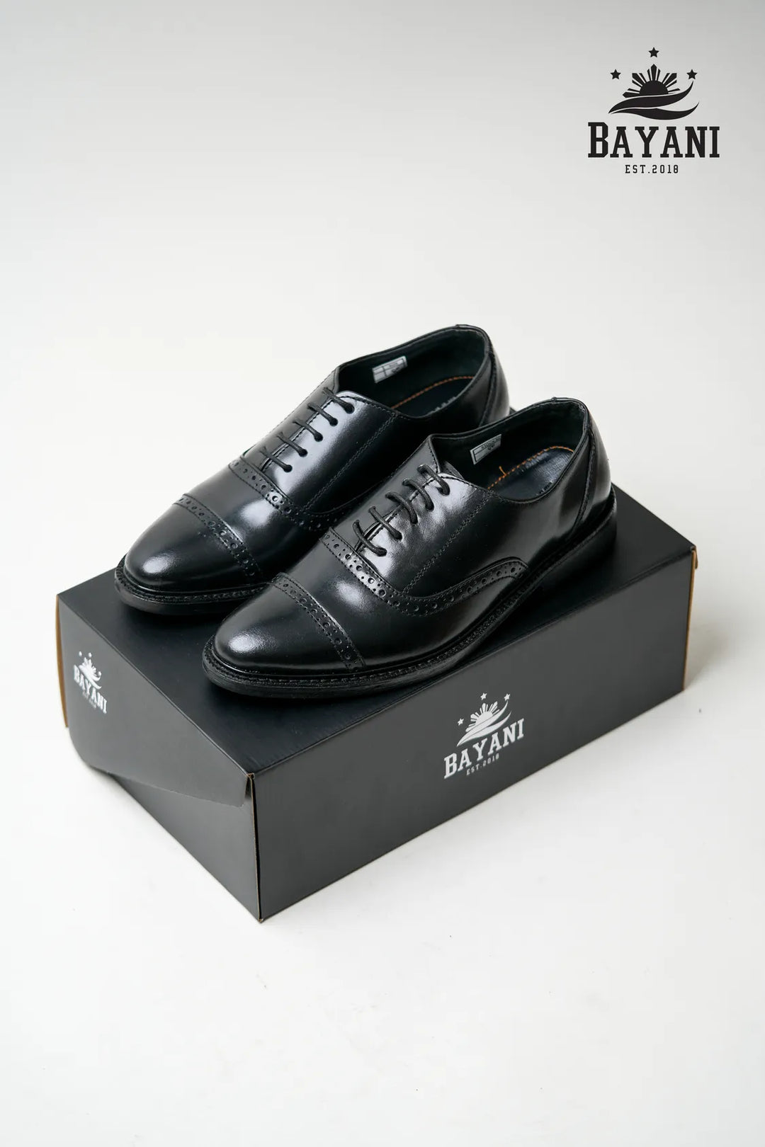 Black dress shoes on a black box with Bayani logo above, on a white background
