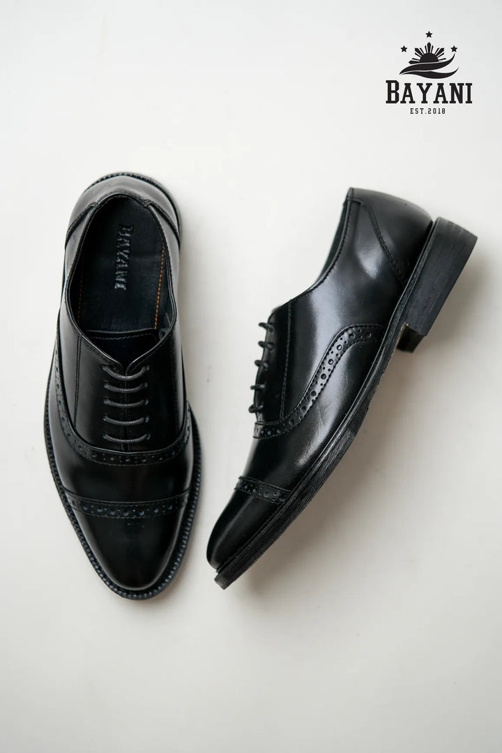 Pair of black dress shoes on a white background with Bayani logo.
