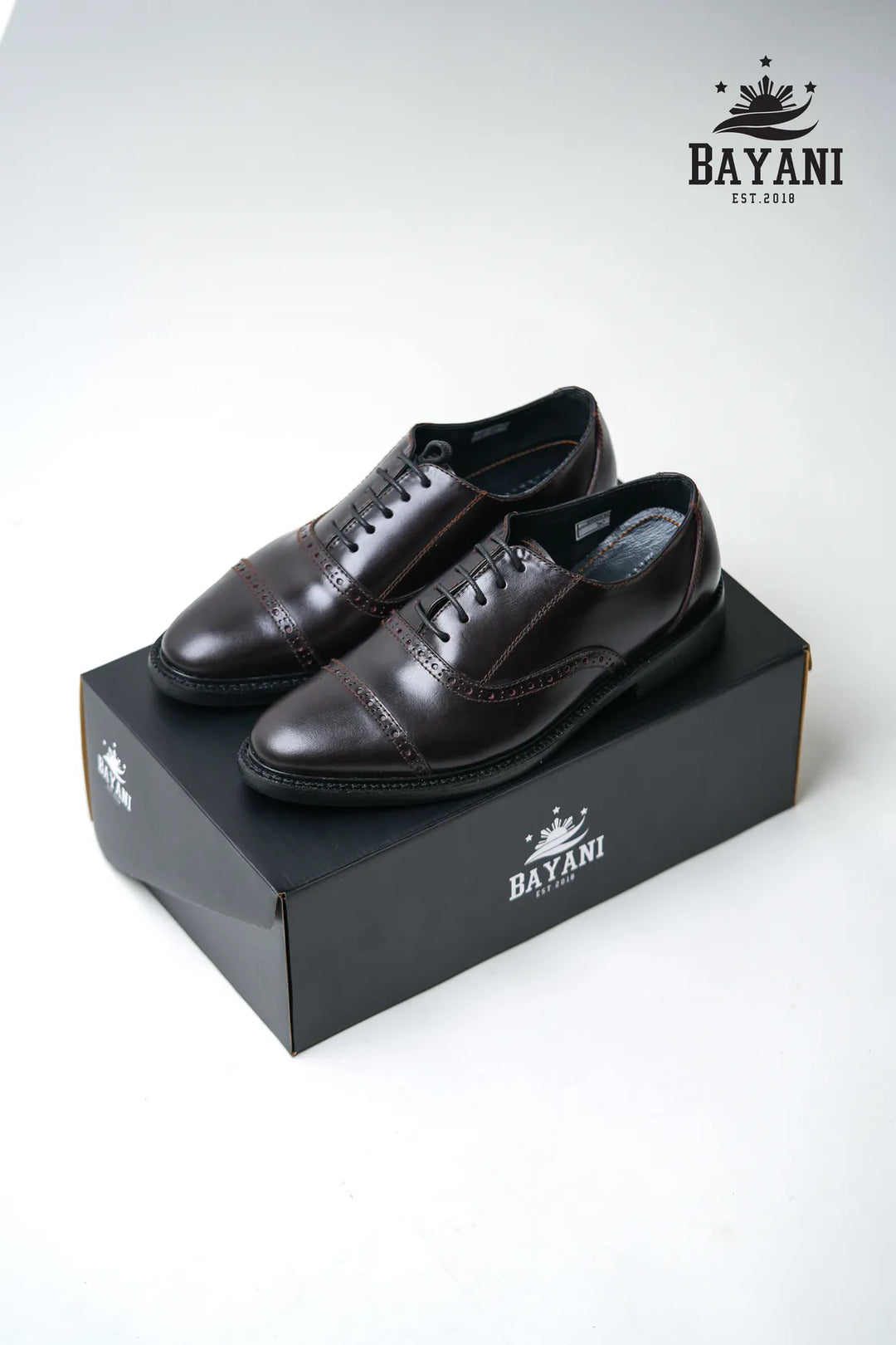 Pair of black dress shoes on a Bayani branded box with the Bayani logo above.

