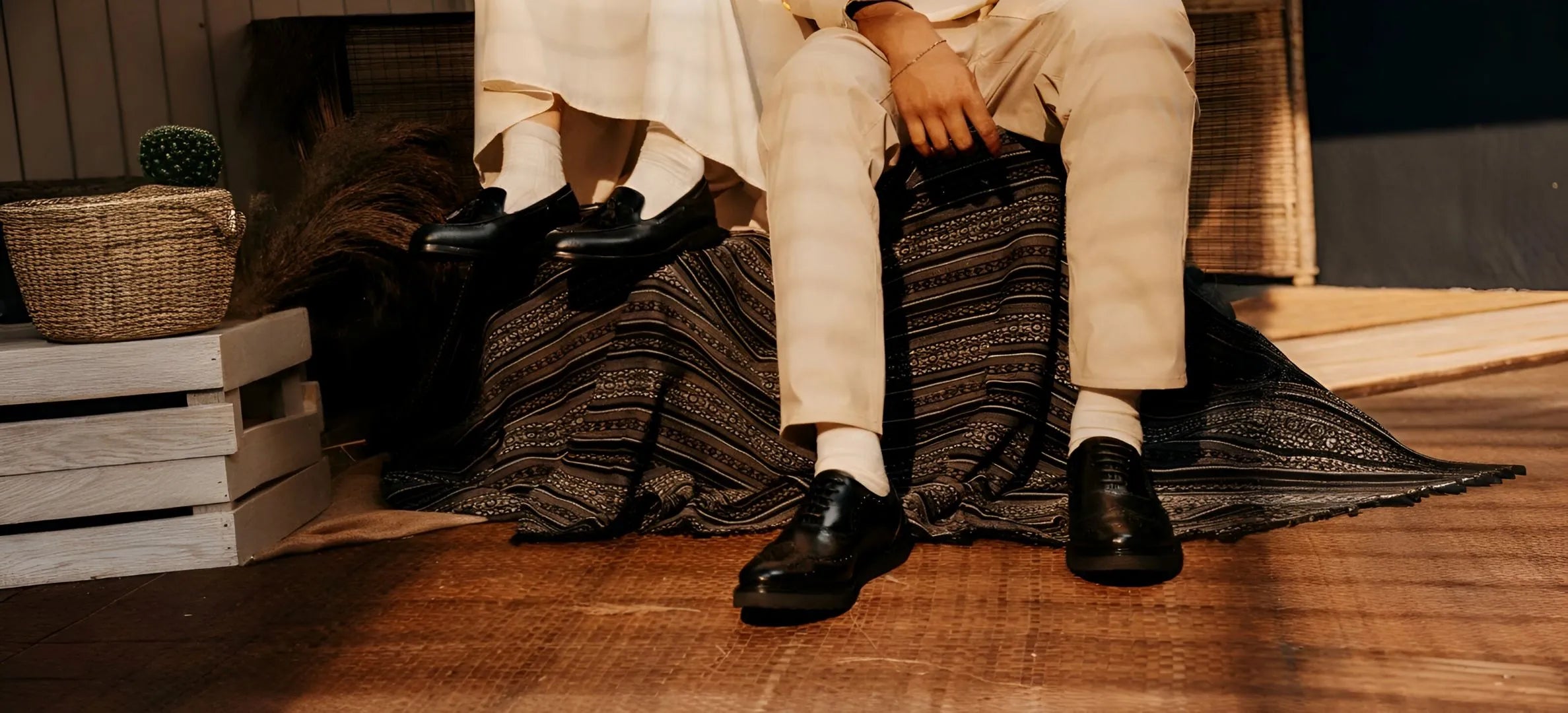 Person sitting on a woven chair with patterned cushions, wearing white pants and black shoes.