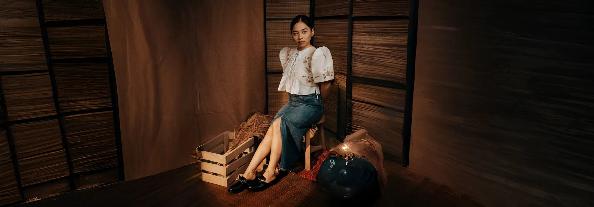 Woman wearing a pair of black shoes sitting on a chair with a wooden crate and bag in a dimly lit room.