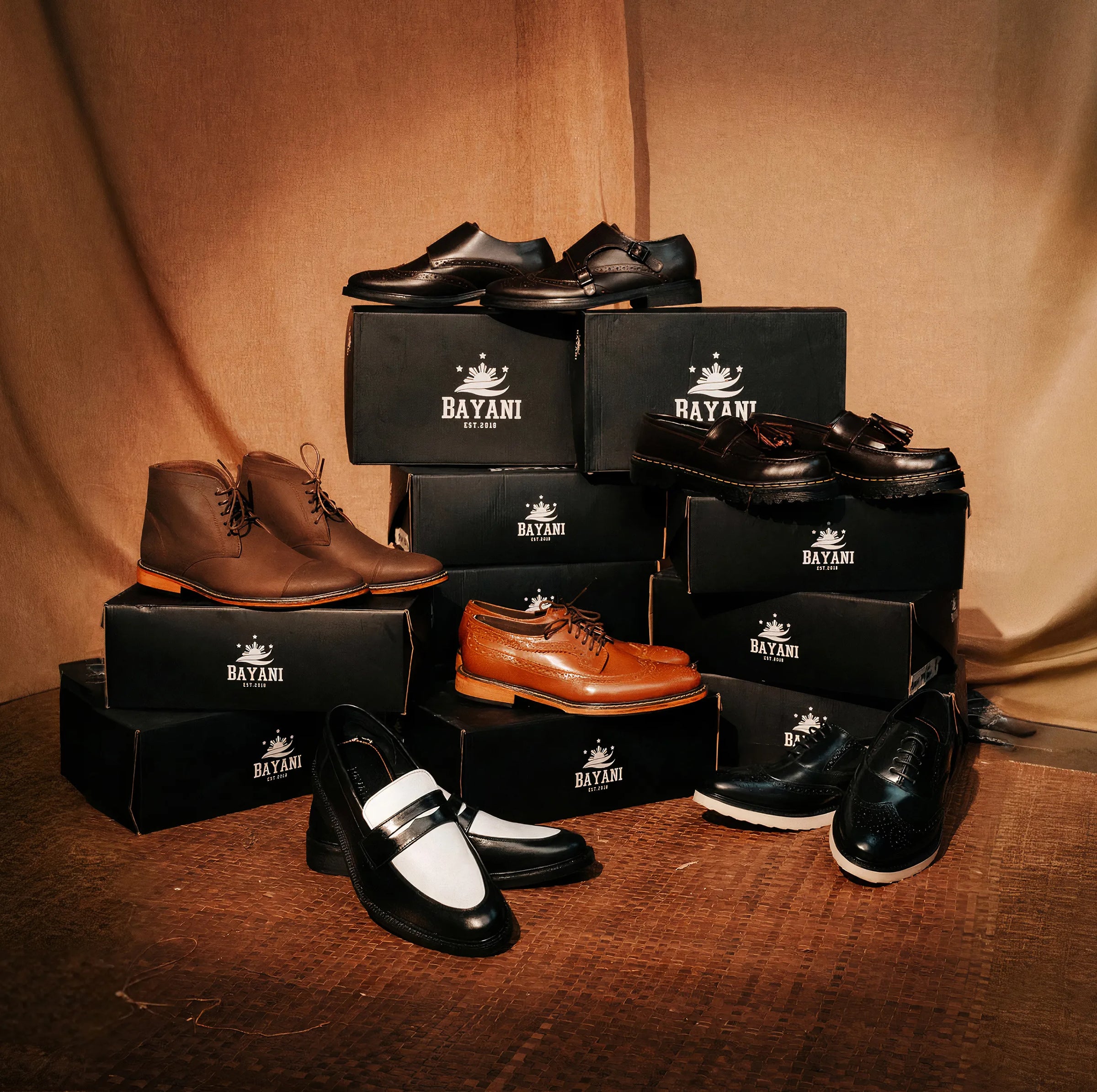 Collection of men's shoes displayed on black boxes with 'BAYANI' branding against a brown fabric background.