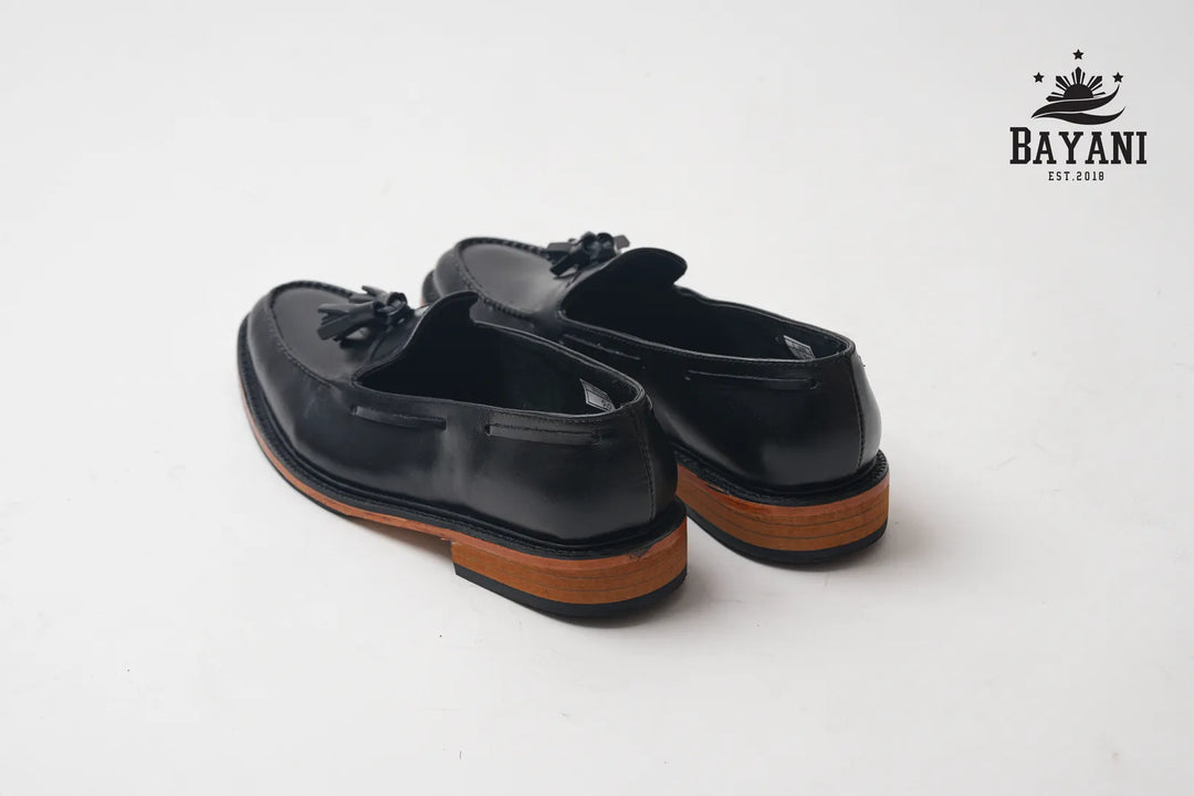 Black loafers with brown soles on a white background, featuring the Bayani logo.
