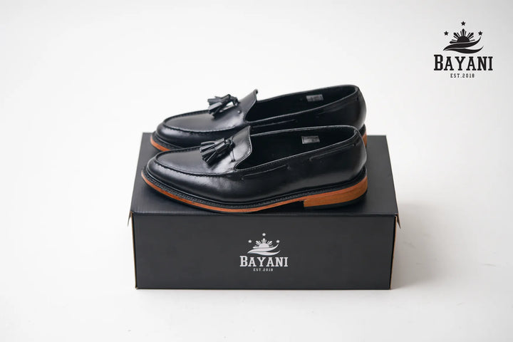 Black loafers with tassels on a black box, featuring the Bayani logo.

