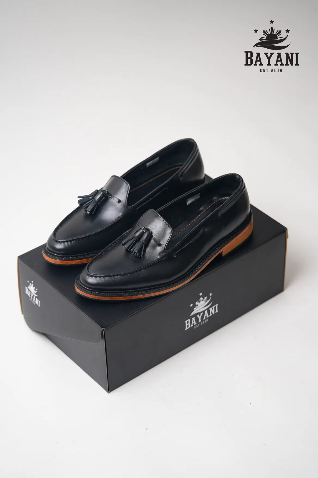 Black loafers with tassels on a black box, featuring the Bayani brand logo.
