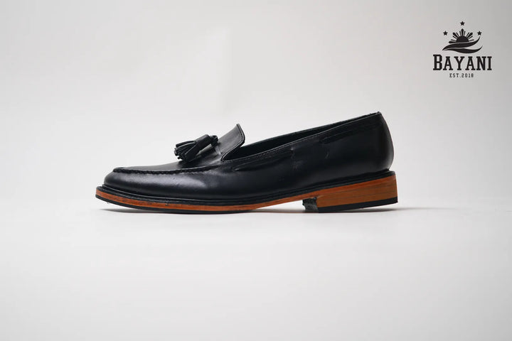 Black loafer shoe with a brown sole on a white background, featuring the Bayani brand logo.
