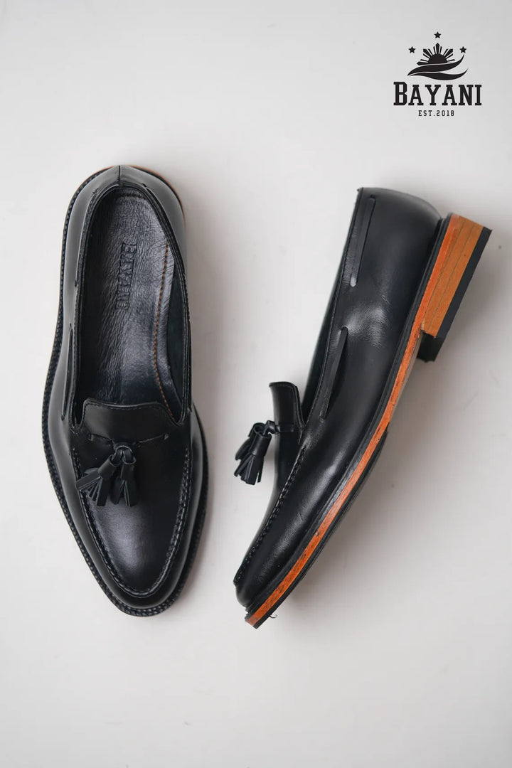 Black loafers with brown soles on a white background, featuring the Bayani brand logo.
