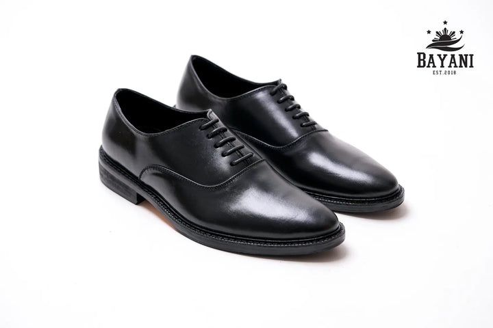 Pair of black dress shoes on a white background with Bayani logo.
