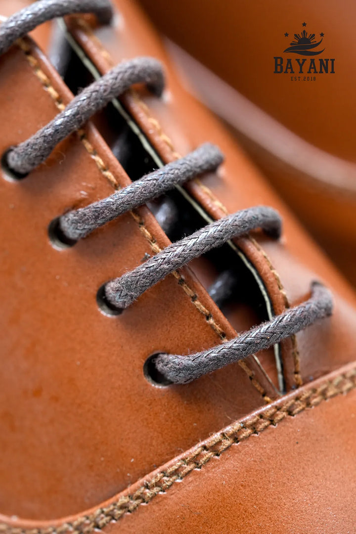 Close-up of a brown leather shoe with gray laces, featuring the Bayani logo.
