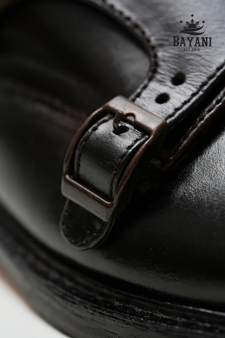 Close-up of a black leather sandal with a strap, featuring the Bayani brand logo.
