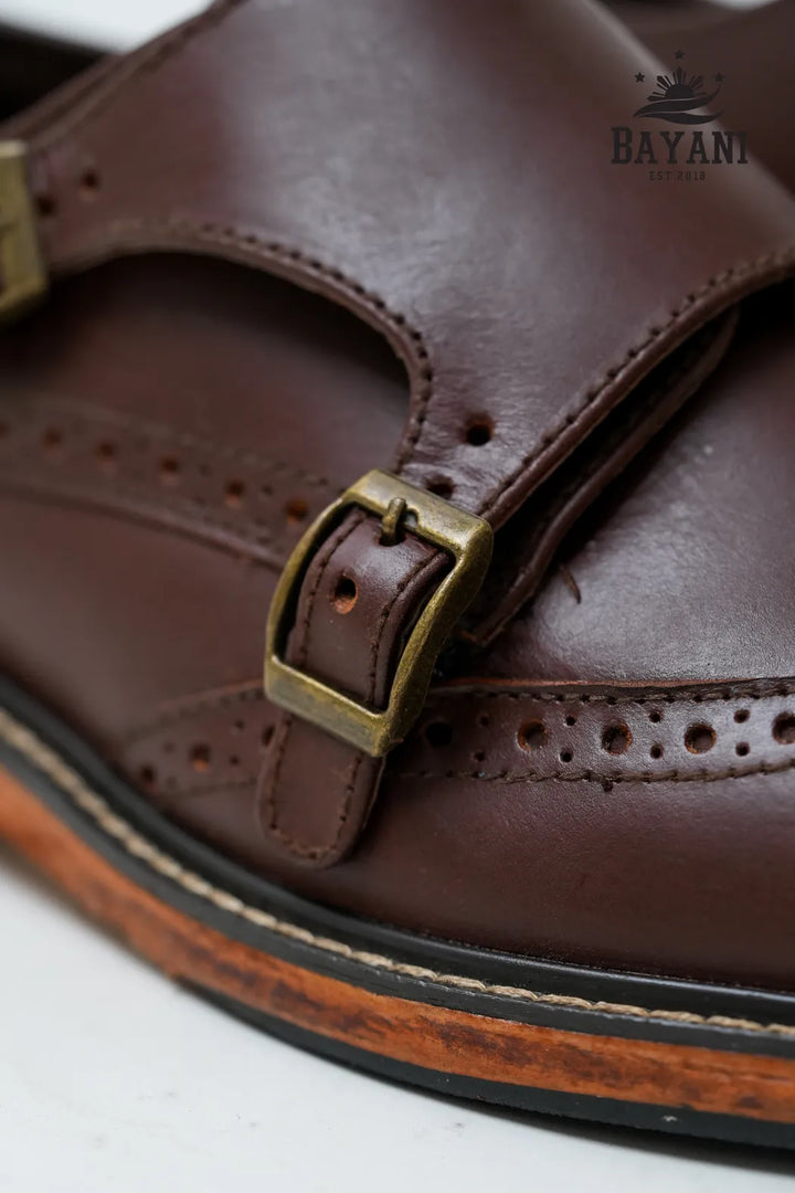 Close-up of a brown leather sandal with a gold buckle on a white background, featuring the Bayani brand.
