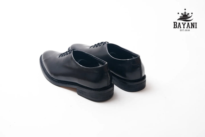 Pair of black dress shoes on a white background with Bayani logo.
