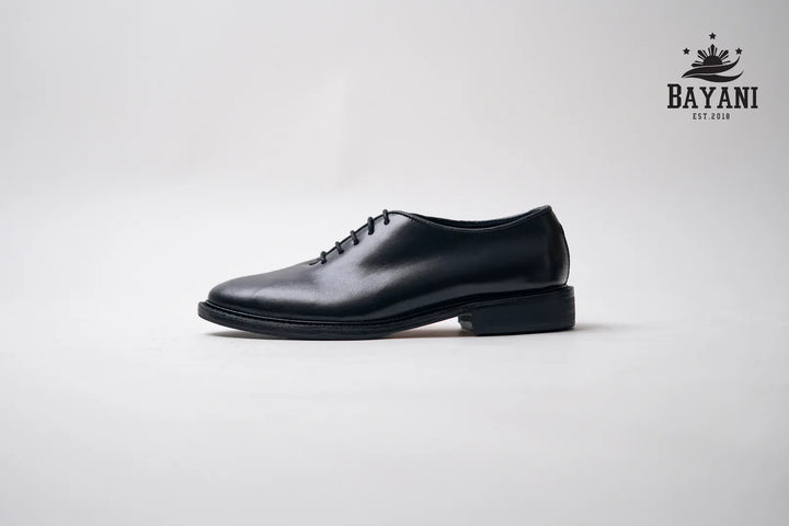 Black leather shoe on a light gray background with Bayani logo.
