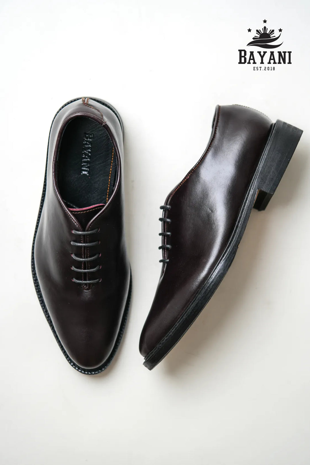 Pair of black dress shoes on a white background with Bayani logo.

