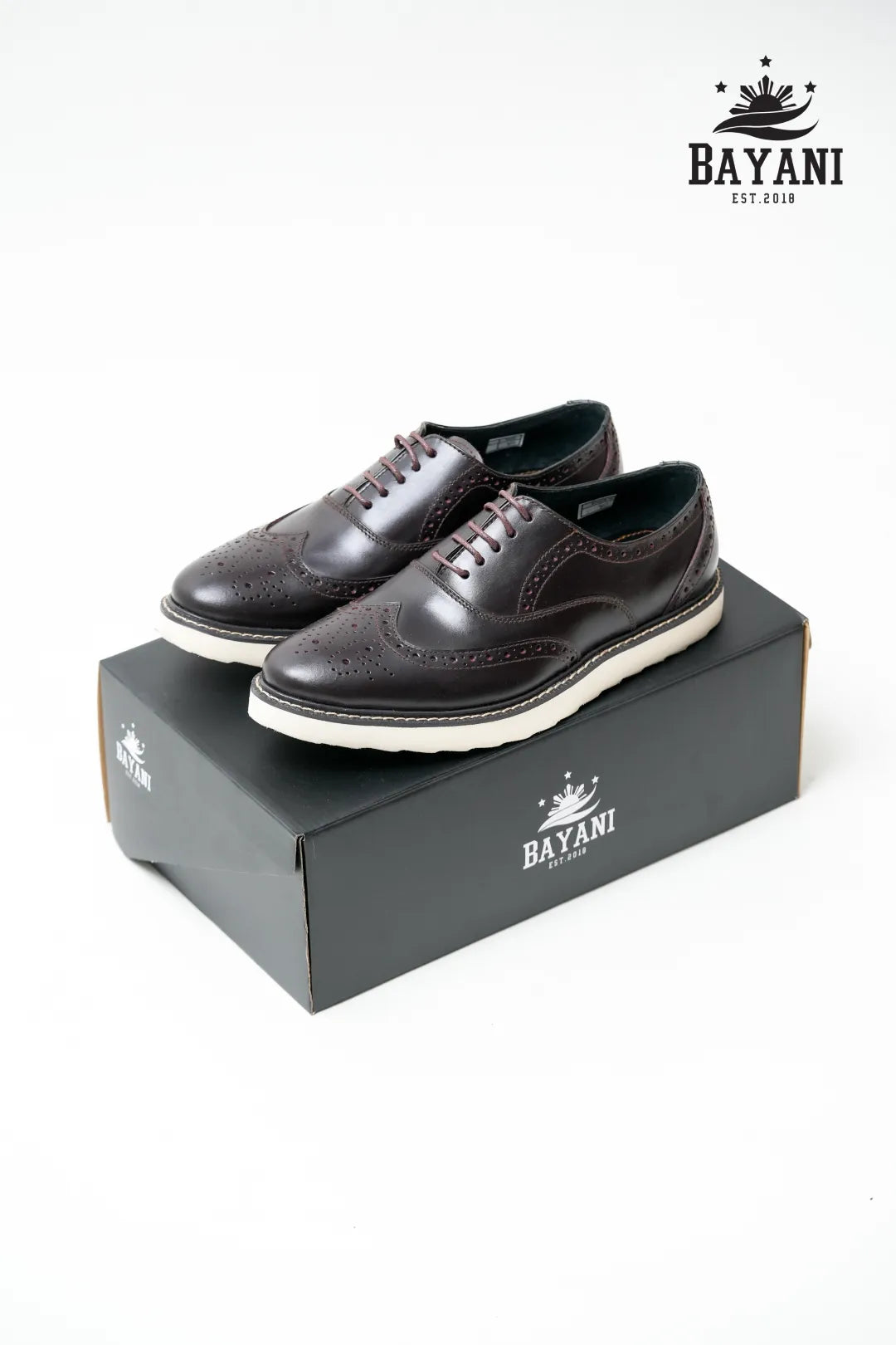 Brown leather shoes with white soles on a Bayani branded box against a white background
