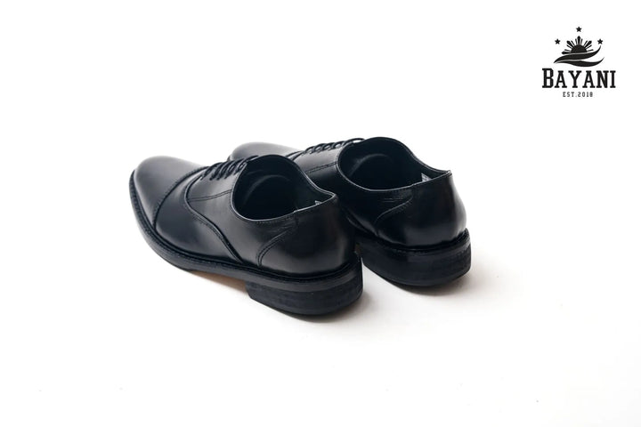 Pair of black dress shoes on a white background with Bayani logo.
