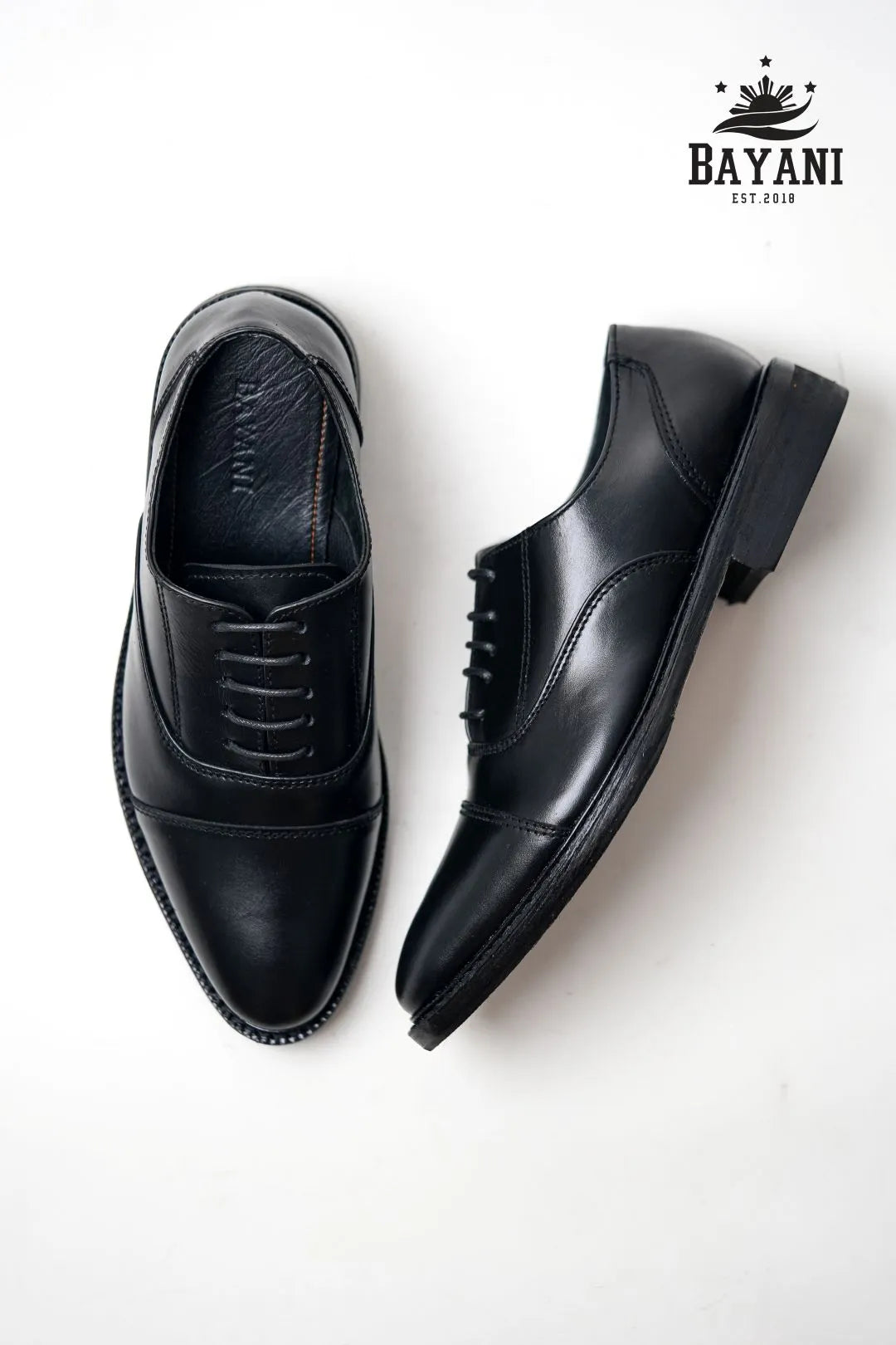 Black leather shoes on a white background with Bayani logo.
