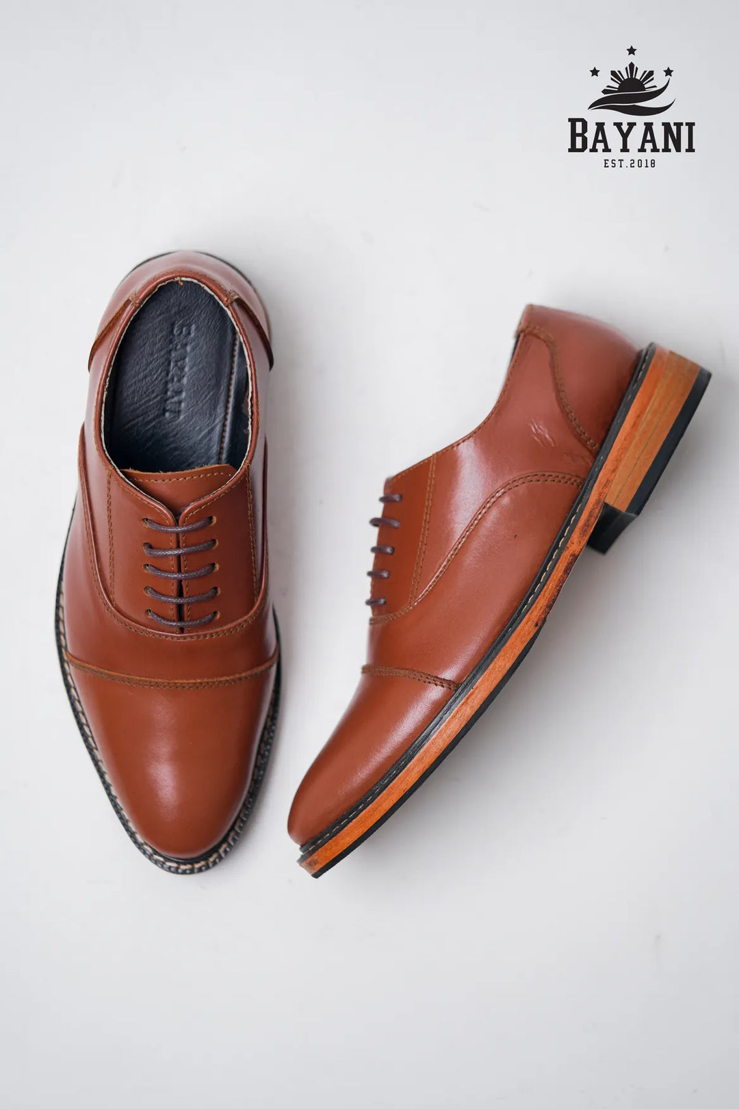 Pair of brown leather shoes on a white background with Bayani logo.
