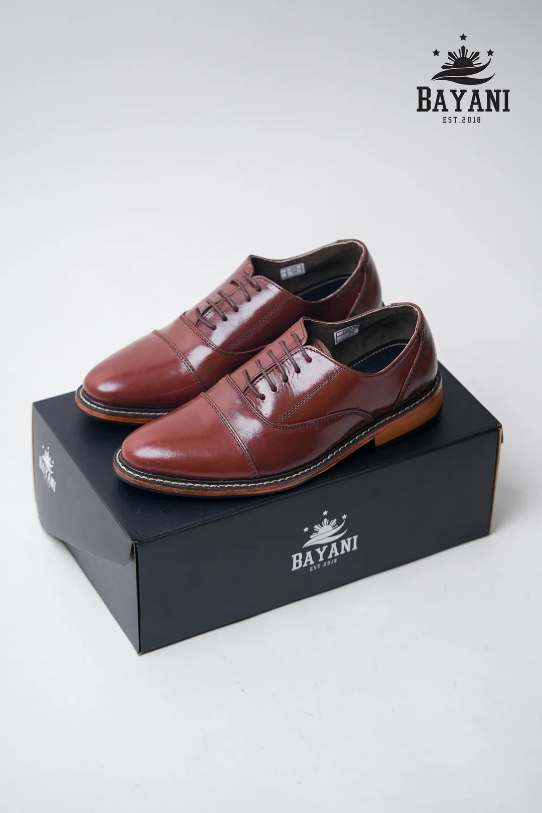 Brown leather shoes on a black box with Bayani logo, on a white background
